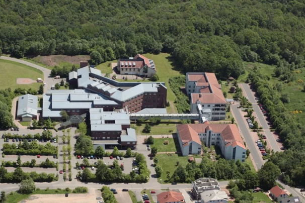 Luftaufnahme eines großen Gebäudekomplexes mit mehreren verbundenen Flügeln, umgeben von Parkplätzen und Wald