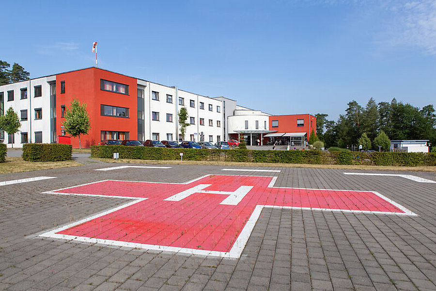 Großes rotes Kreuz mit weißem A auf gepflastertem Vorplatz vor einem mehrstöckigen Gebäude mit roten und weißen Fassadenteilen und Bäumen im Hintergrund