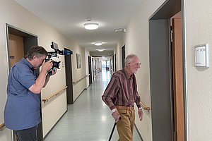 Ein älterer Mann mit Gehstock betritt einen Flurraum, während ein Kameramann ihn filmt.