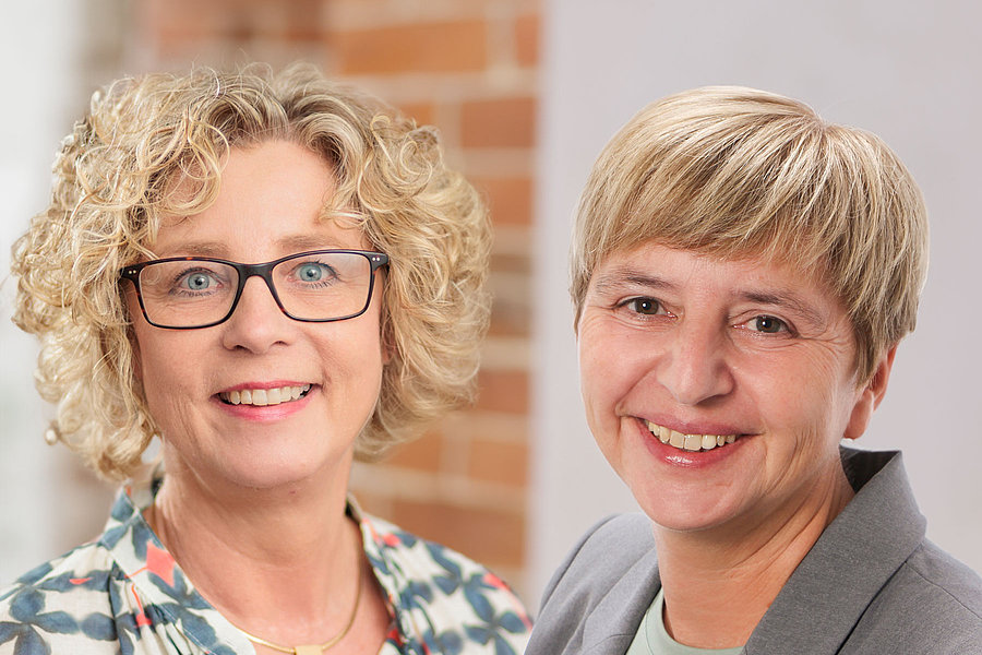 Oberin Helga Schmitt mit lockigem Haar und gemustertem Oberteil neben Kaufmännischer Direktorin Natalie Hirschauer mit kurzem blonden Haar und grauem Blazer vor Backsteinwand
