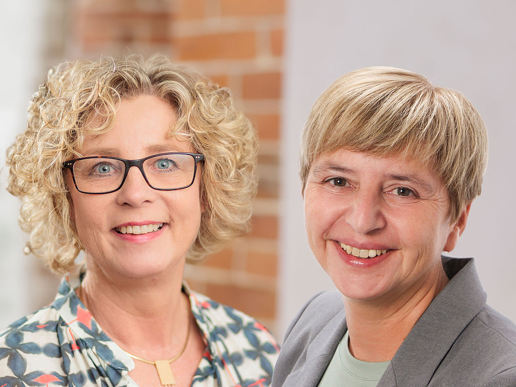 Oberin Helga Schmitt mit lockigem Haar und gemustertem Oberteil neben Kaufmännischer Direktorin Natalie Hirschauer mit kurzem blonden Haar und grauem Blazer vor Backsteinwand