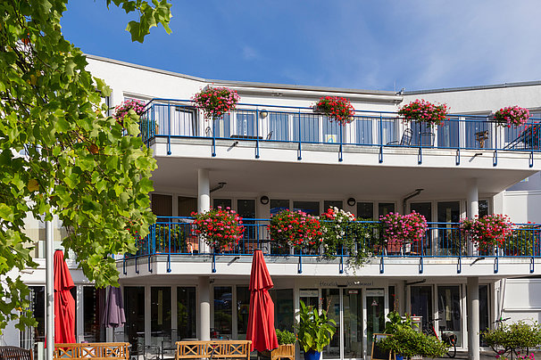 Mehrstöckiges Gebäude mit Balkonen, die mit roten Blumen in Blumenkästen geschmückt sind, und mehreren Sonnenschirmen im Vordergrund.