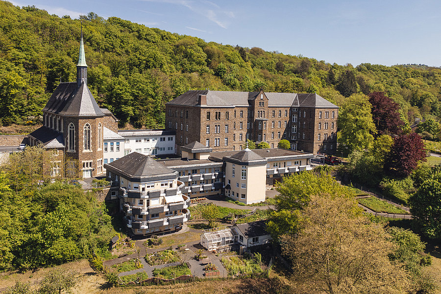 Luftaufnahme eines historischen Gebäudes mit Kirche und modernen Anbauten, umgeben von Wald.