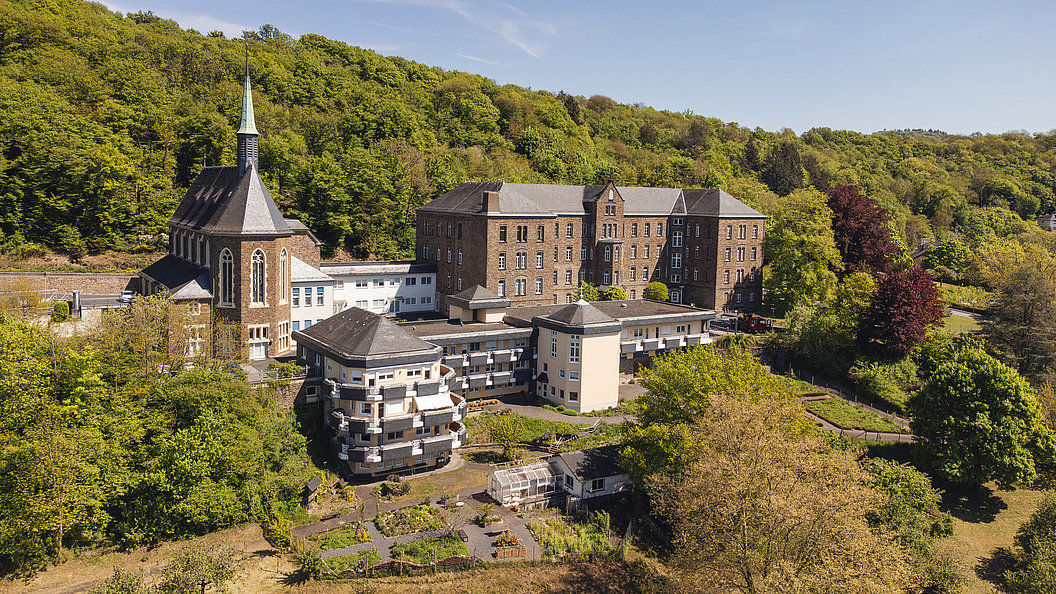 Luftaufnahme eines historischen Gebäudes mit Kirche und modernen Anbauten, umgeben von Wald.