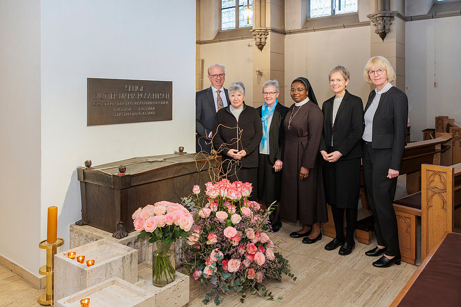 Fünf Personen stehen in einer Kirche neben einem großen Blumenarrangement aus rosa Rosen und einem Kerzenständer mit brennenden Kerzen