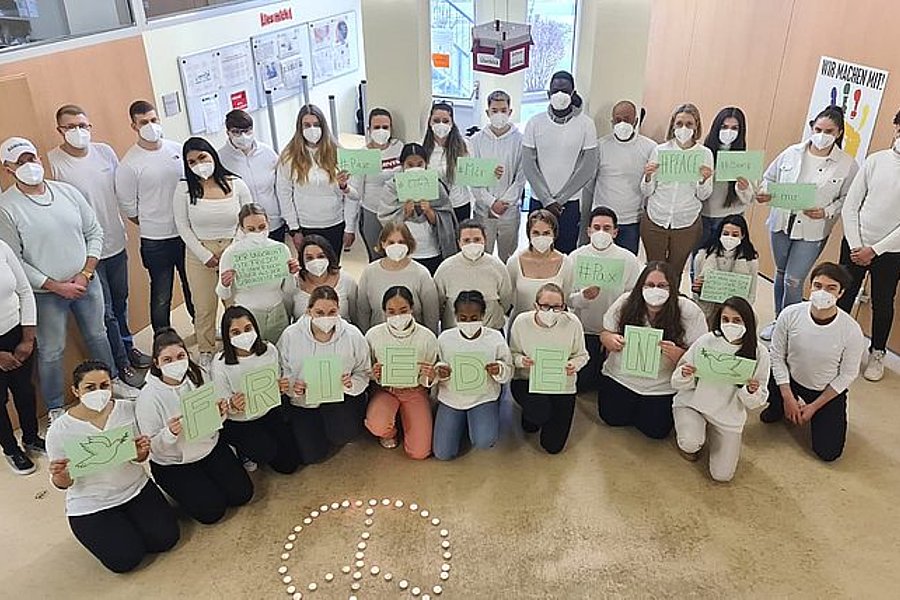 Gruppenfoto von 29 Personen mit weißen Masken und weißen Oberteilen, die grüne Schilder mit Buchstaben halten, die zusammen das Wort 'FREIHEIT' bilden, vor einem Friedenszeichen aus Kerzen auf dem Boden
