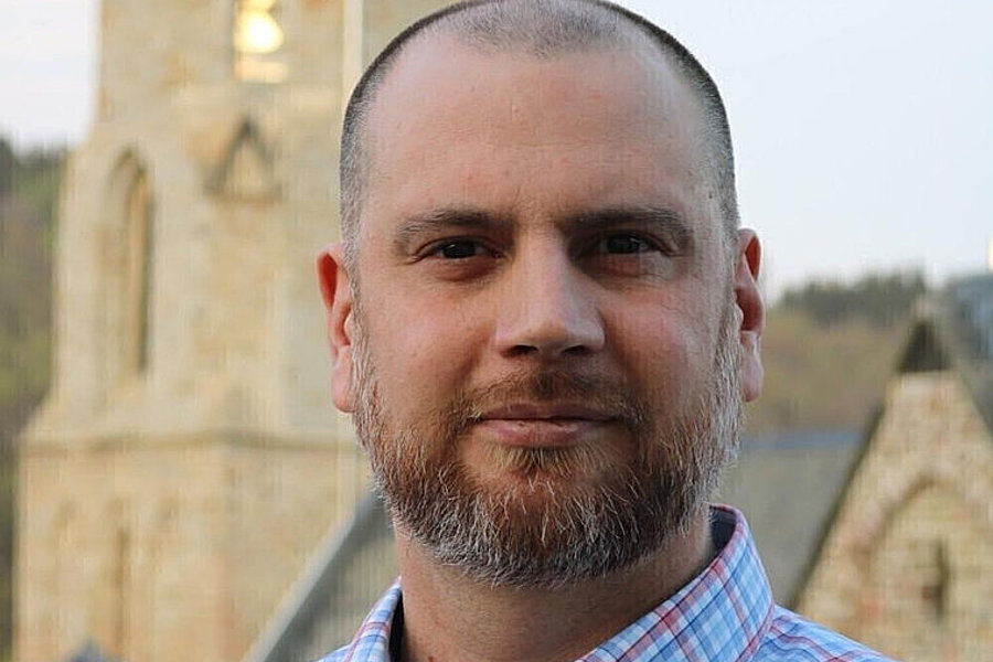 Mann mit kurzem Bart und kariertem Hemd vor einer historischen Kirche mit Turm