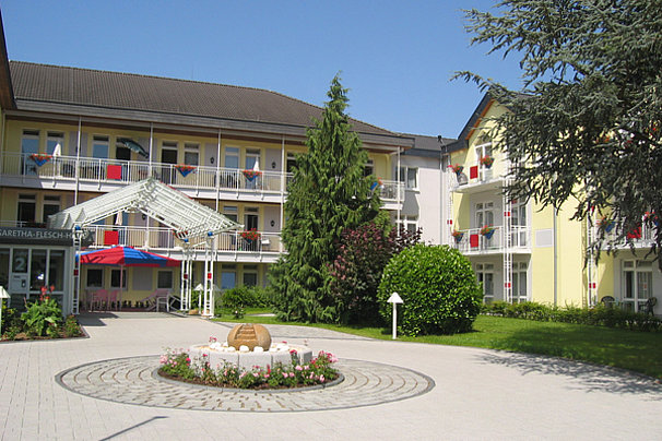 Innenhof eines mehrstöckigen Gebäudes mit Balkon, zentralem Brunnen und bepflanztem Gartenbereich.