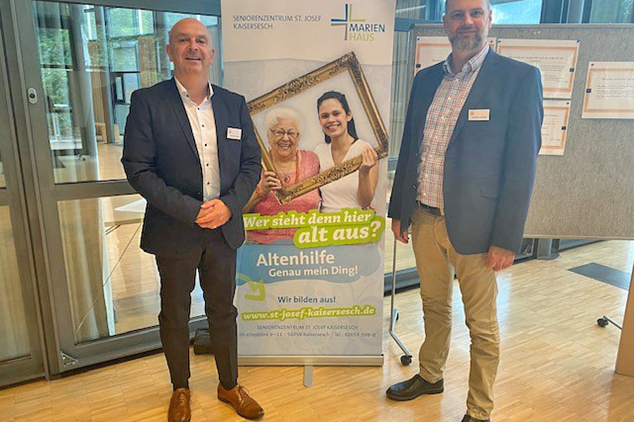 Zwei Männer in Anzügen stehen neben einem Banner des Seniorenzentrums St. Josef Kaisersesch mit einem Bild von zwei Frauen, eine ältere und eine jüngere, die einen Bilderrahmen halten. Das Banner wirbt für Altenhilfe und Ausbildung.