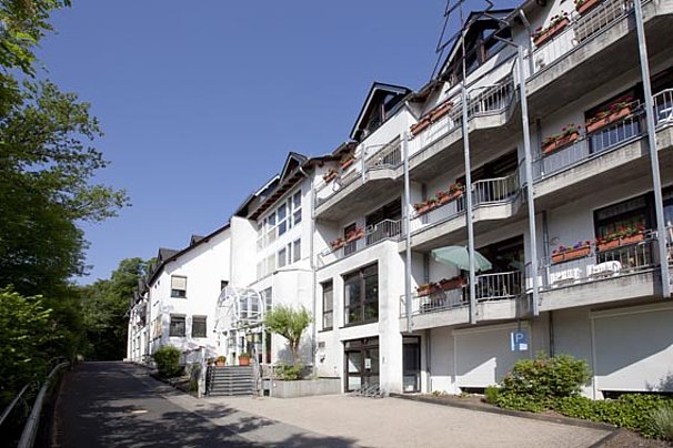 Mehrstöckiges Wohngebäude mit Balkonen, Blumenkästen und einer Einfahrt, umgeben von Bäumen und einem Gehweg.