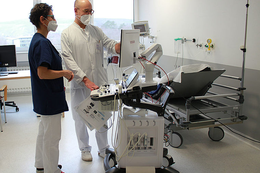 Zwei medizinische Fachkräfte mit Masken bedienen ein mobiles Ultraschallgerät in einem Krankenhauszimmer mit leerem Patientenbett