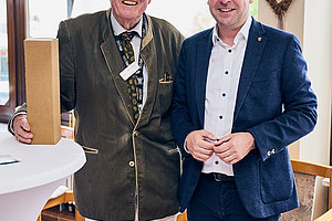 Zwei Männer in einem hellen Raum, der eine trägt eine grüne Jacke und hält eine Holzbox, der andere einen blauen Anzug und ein weißes Hemd