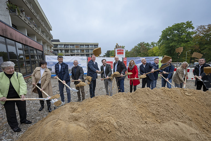 Mehrere Personen stehen nebeneinander und halten Schaufeln, mit denen sie Sand aus einer großen Sandhaufen schaufeln.