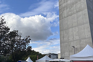 Hohes graues Gebäude mit Schriftzug 'MARIENHAUS' neben mehreren weißen Zelten und Menschen auf einem Platz unter blauem Himmel mit Wolken.