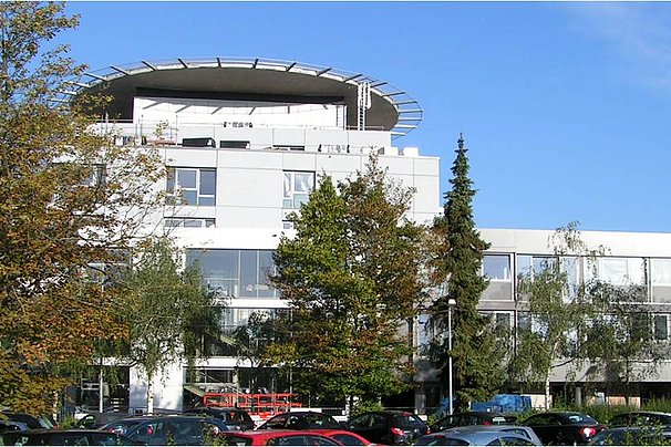 Modernes Bürogebäude mit mehreren Stockwerken, umgeben von Bäumen und parkenden Autos unter blauem Himmel