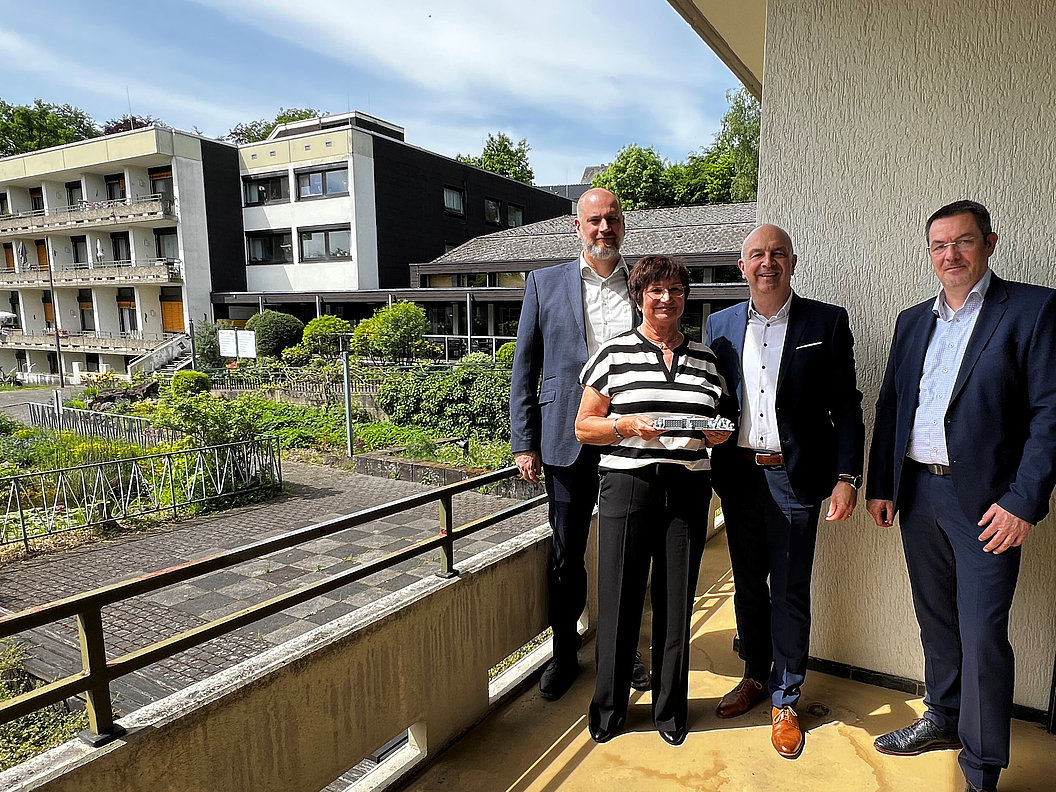 Vier Personen in formeller Kleidung stehen auf einem Balkon mit Blick auf ein mehrstöckiges Gebäude und einen Gartenbereich.