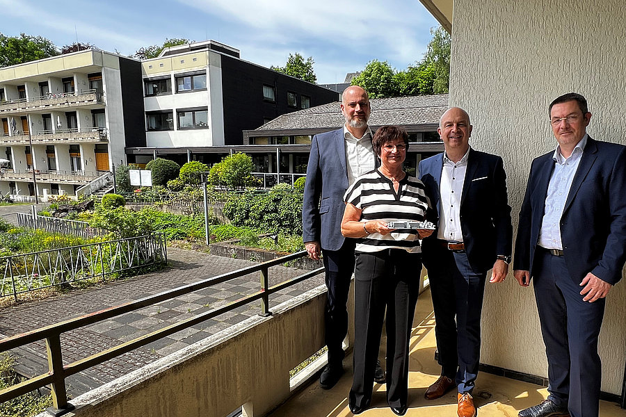 Vier Personen in formeller Kleidung stehen auf einem Balkon mit Blick auf ein mehrstöckiges Gebäude und einen Gartenbereich.