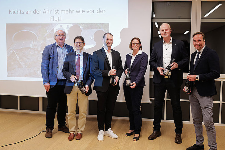 Fünf Personen in formeller Kleidung stehen vor einer Präsentationsleinwand mit dem Text 'Nichts an der Ahr ist mehr wie vor der Flut!' und halten Weinflaschen in der Hand.