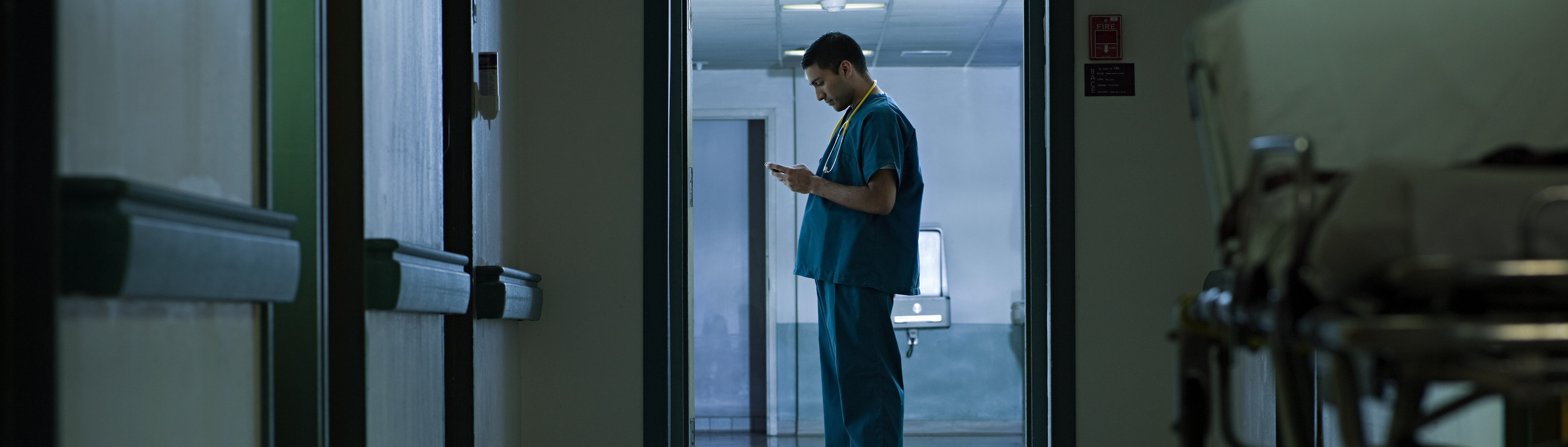 Krankenhausflur mit einem stehenden Mann in medizinischer Kleidung, der auf ein Tablet schaut, im Vordergrund ein leerer Rollstuhl