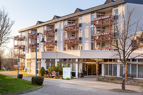 Mehrstöckiges Gebäude mit mehreren Balkonen, Eingangsbereich mit Glasfront und Schildern, umgeben von Bäumen und Gehwegen.