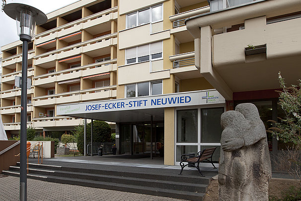Mehrstöckiges Gebäude mit Balkonen und dem Schriftzug 'Josef-Ecker-Stift Neuwied' über dem Eingang, davor eine Steinskulptur zweier umarmender Figuren.