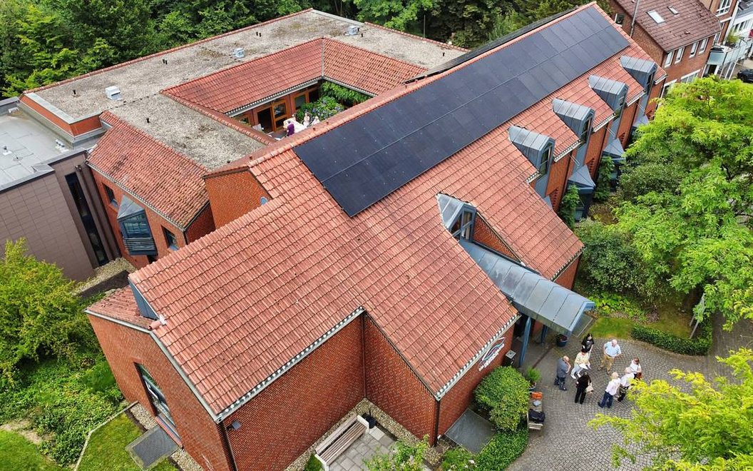 Luftaufnahme eines roten Backsteingebäudes mit Solarpaneelen auf dem Dach und mehreren Personen vor dem Eingang.