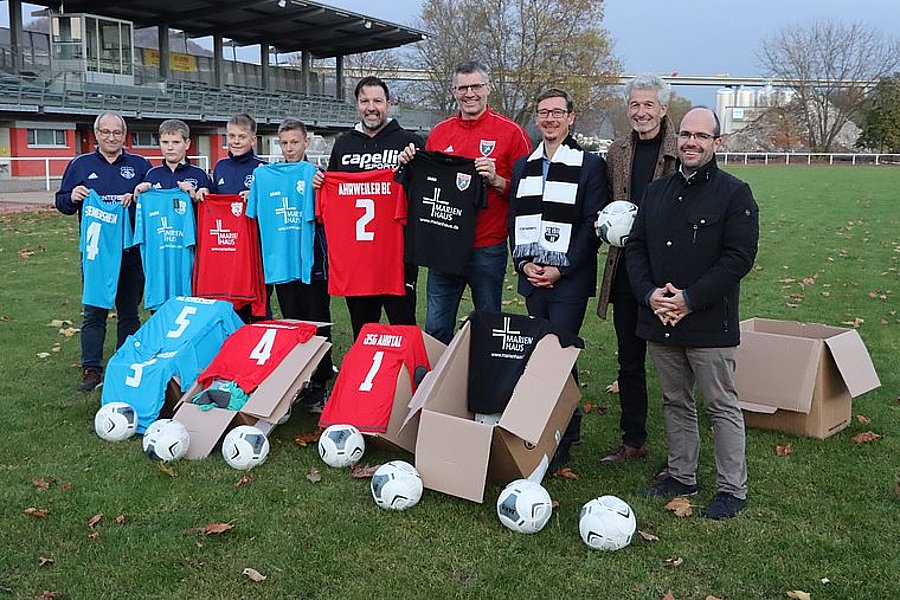 Gruppe von neun Personen auf einem Fußballfeld mit Fußballtrikots und Bällen, einige halten Trikots in Hellblau, Rot oder Schwarz