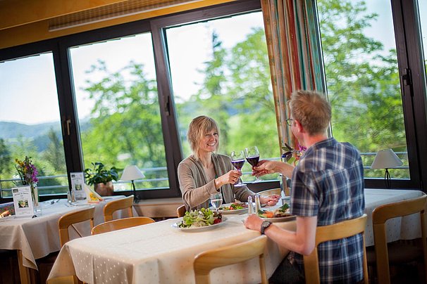 Paar stößt mit Rotweingläsern in einem Restaurant mit großen Fenstern und Blick auf grüne Bäume an