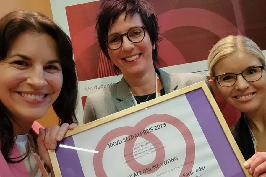 Drei Frauen halten eine Urkunde für den 1. Platz des kkvd Sozialpreises 2023, lächelnd vor einem Banner mit dem kkvd Sozialpreis-Logo