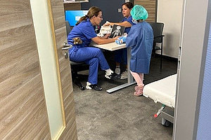 Zwei medizinische Fachkräfte in blauer Kleidung sitzen an einem Tisch, eine dritte Person in OP-Haube und Kittel steht daneben in einem Behandlungszimmer.