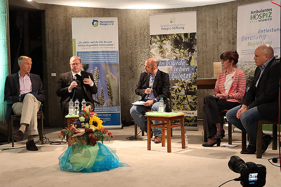 Fünf Personen sitzen auf einer Bühne bei einer Podiumsdiskussion vor Bannern von Hospizorganisationen, eine Person spricht in ein Mikrofon.