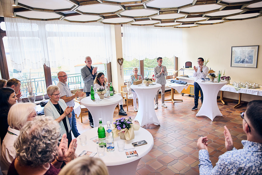 Gruppe von Menschen steht und sitzt in einem Raum mit Tischen und applaudiert