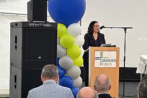 Frau am Rednerpult mit Mikrofon vor Luftballon-Säule in Blau, Weiß, Hellgrün und Grau, davor sitzende Zuhörer, Lautsprecher links.