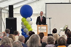 Mann im Anzug steht am Rednerpult mit Mikrofon vor Publikum in Zelt, neben ihm Säule aus blauen, weißen und grünen Luftballons, im Hintergrund Leinwand.