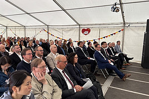 Menschen in formeller Kleidung sitzen in einem Zelt auf Stühlen und hören einer Veranstaltung zu.
