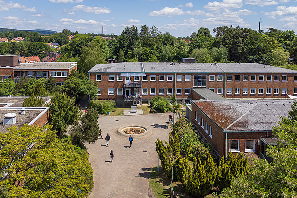 Schulgebäude aus Backstein mit Innenhof und einigen Personen, umgeben von Bäumen und grüner Landschaft unter blauem Himmel mit Wolken