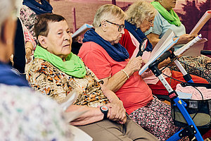 Gruppe älterer Menschen in bunten Schals sitzt auf Stühlen und liest Liederblätter.