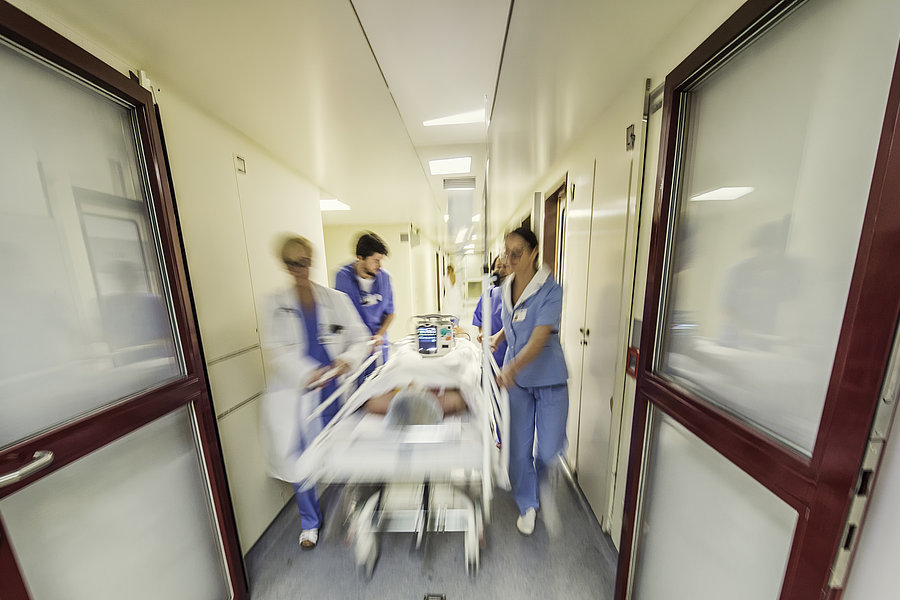 Krankenhausflur mit drei medizinischem Personal in blauer Schutzkleidung, die eine liegende Person auf einer Trage eilen