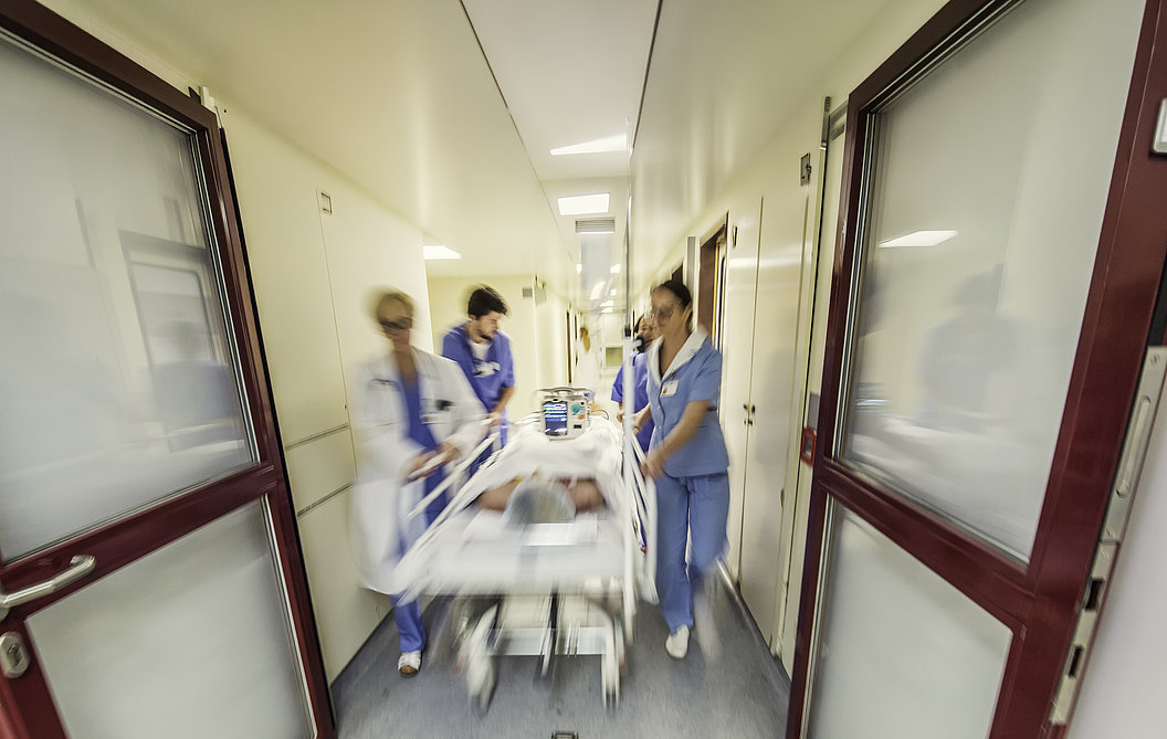 Krankenhausflur mit drei medizinischem Personal in blauer Schutzkleidung, die eine liegende Person auf einer Trage eilen
