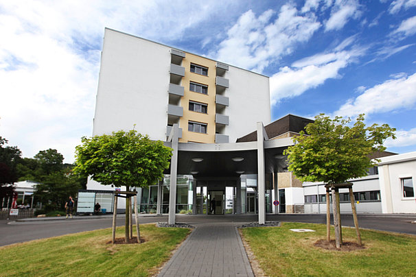 Moderne Gebäude mit sechs Stockwerken, Eingangsbereich mit Glasfront und zwei Bäumen auf der linken und rechten Seite.