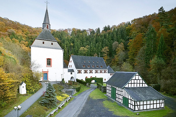 Kloster Ehrenstein mit weißem Kirchturm, Fachwerkhaus und Torbogen, umgeben von herbstlich gefärbtem Wald