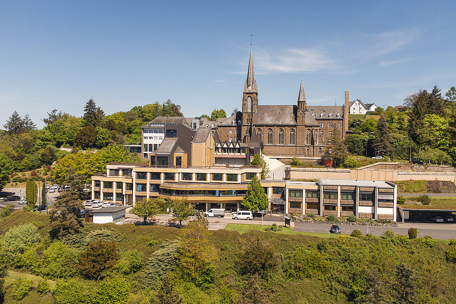 Blick auf ein mehrstöckiges Gebäude mit flachem Dach vor einer Kirche mit zwei Türmen, umgeben von Bäumen und Grünflächen.