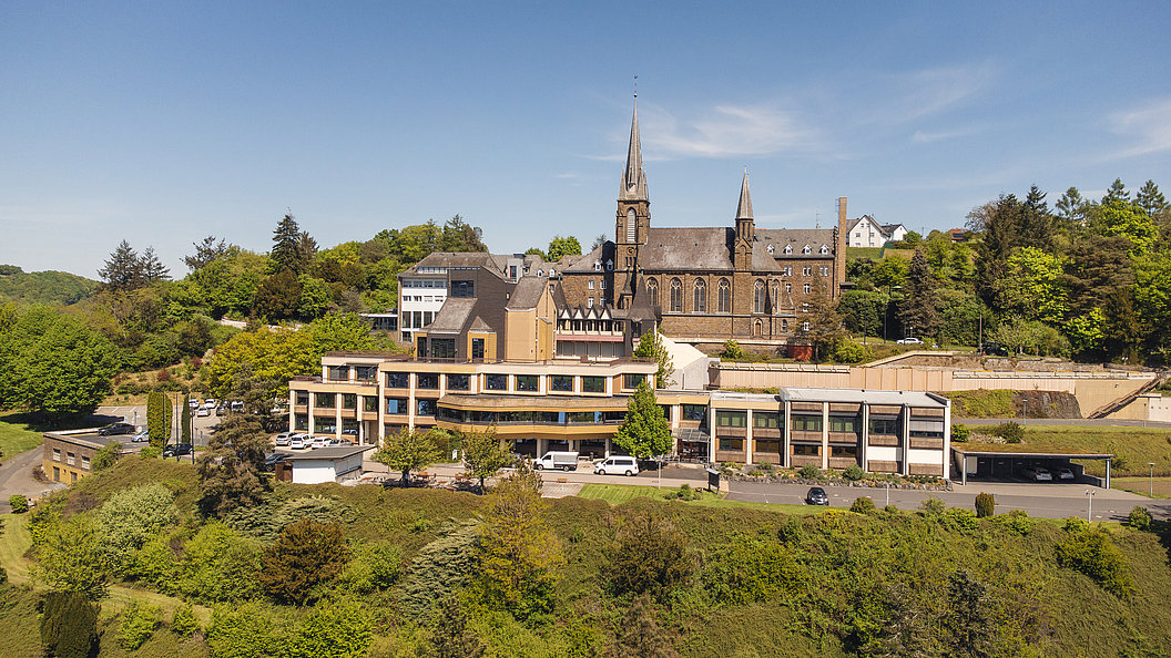 Blick auf ein mehrstöckiges Gebäude mit flachem Dach vor einer Kirche mit zwei Türmen, umgeben von Bäumen und Grünflächen.