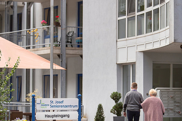 Zwei ältere Personen gehen Hand in Hand auf den Haupteingang eines Seniorenwohnheims mit Balkonen und Pflanzen zu.
