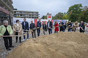 Gruppe von Menschen steht mit Schaufeln vor einem großen Sandhaufen auf einer Baustelle.