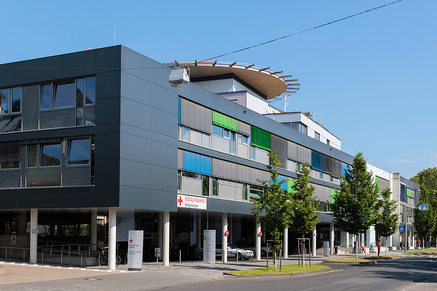 Moderne, mehrstöckige Gebäude mit großen Fenstern und bunten Fassadenelementen. Vor dem Gebäude stehen Bäume und Parkplätze.