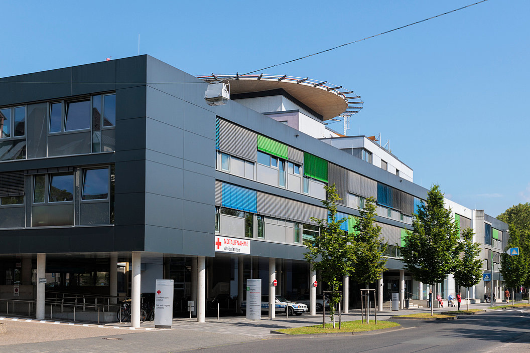 Moderne, mehrstöckige Gebäude mit großen Fenstern und bunten Fassadenelementen. Vor dem Gebäude stehen Bäume und Parkplätze.
