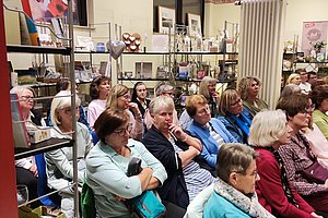Gruppe von Menschen sitzt in einem Raum mit Regalen und Bildern an den Wänden und hört aufmerksam zu
