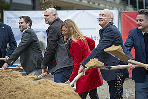Mehrere Personen in formeller Kleidung schaufeln gemeinsam Sand mit Schaufeln.