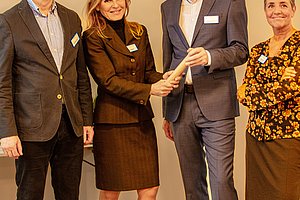 Vier Personen in Businesskleidung stehen nebeneinander, zwei davon schütteln sich die Hände.